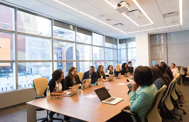 thirteen payroll professionals working together in a large conference room around a wooden table with large glass windows in the background